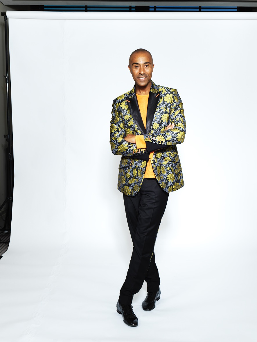 Colin Jackson CBE in a gold and green floral blazer with an orange shirt, arms crossed